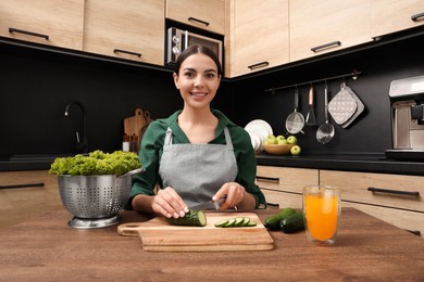 Woman in apron cutting cucumber at wooden table indoors