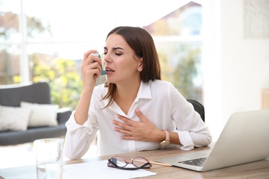 Young woman with asthma inhaler at table in light room