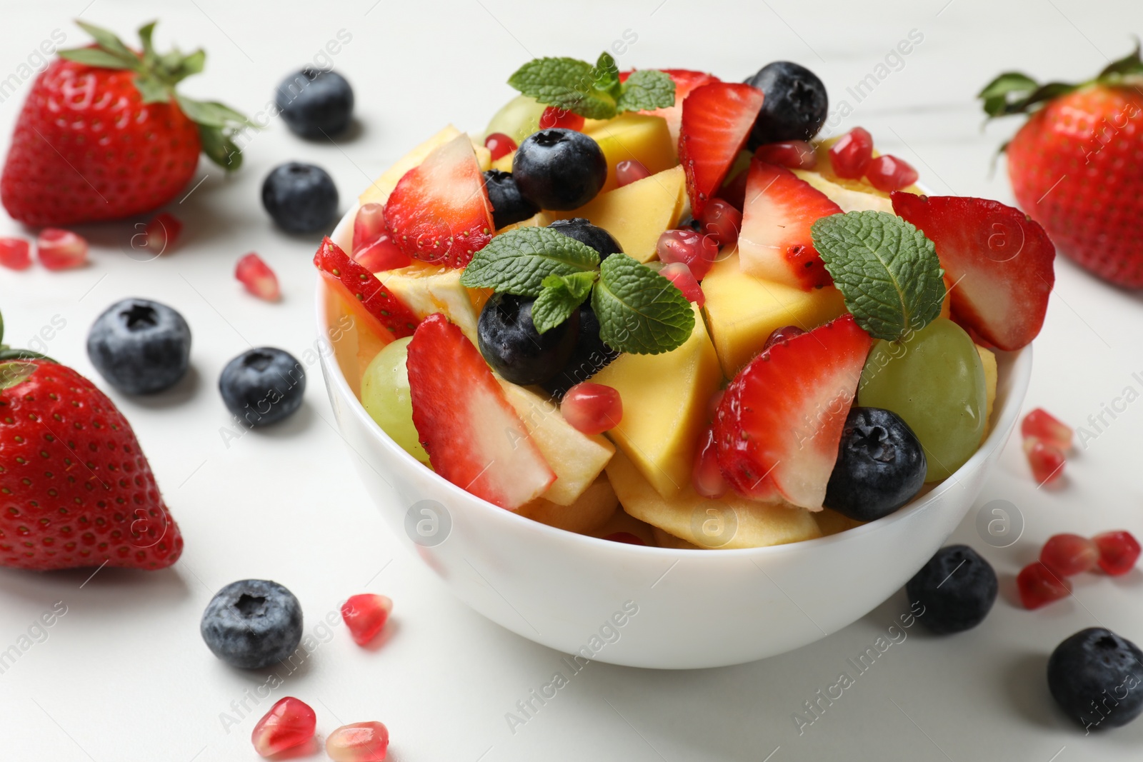 Photo of Tasty fruit salad in bowl and ingredients on white table, closeup