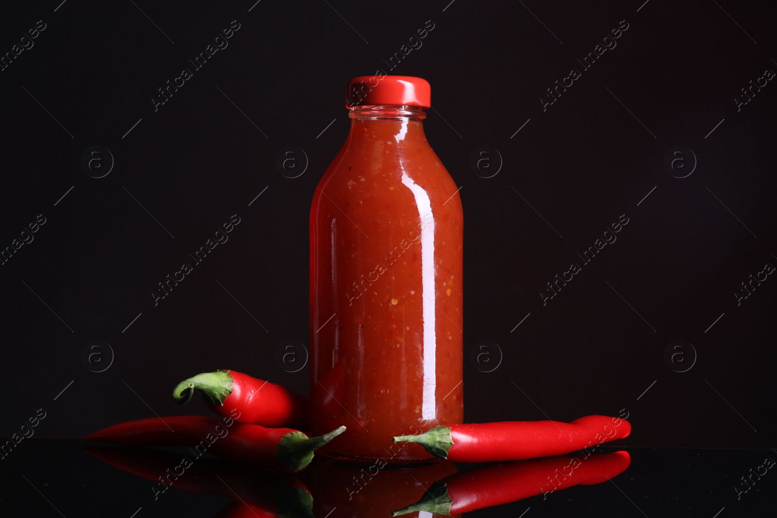 Photo of Spicy chili sauce in bottle and peppers against dark background