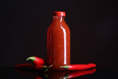 Photo of Spicy chili sauce in bottle and peppers against dark background