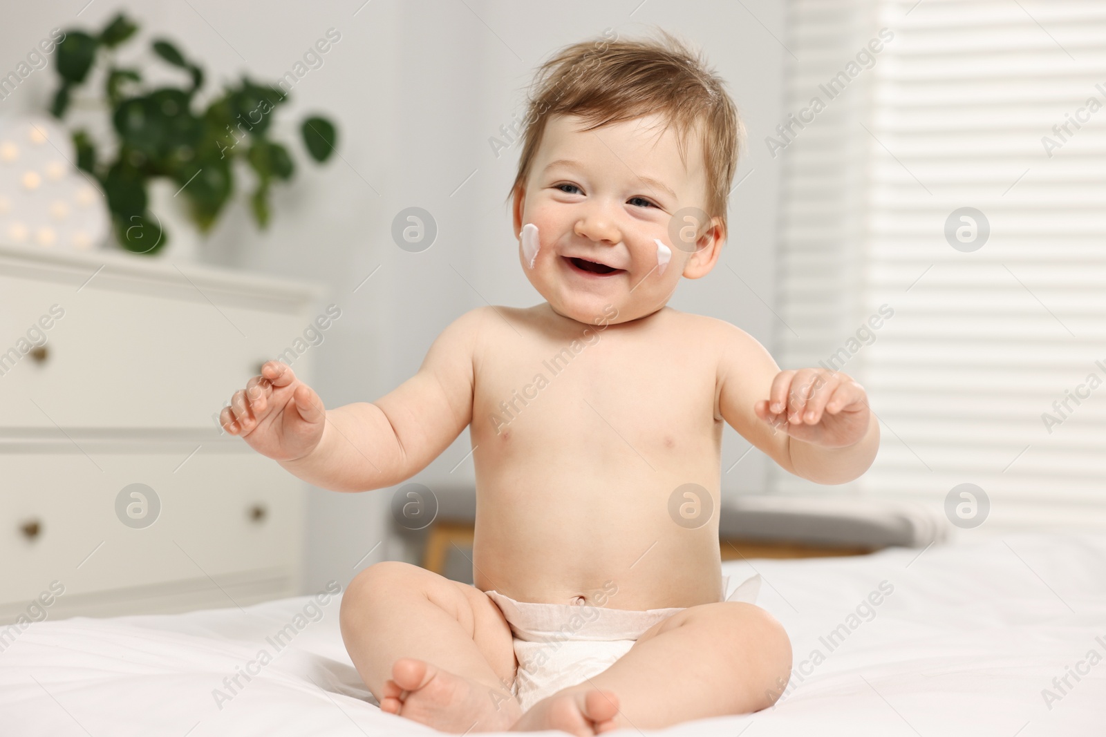 Photo of Cute little baby with moisturizing cream on face sitting on bed at home