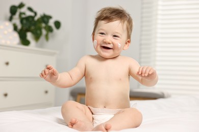 Photo of Cute little baby with moisturizing cream on face sitting on bed at home