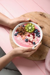 Photo of Woman holding tasty smoothie bowl with fresh kiwi fruit, berries and granola at pink wooden table, closeup