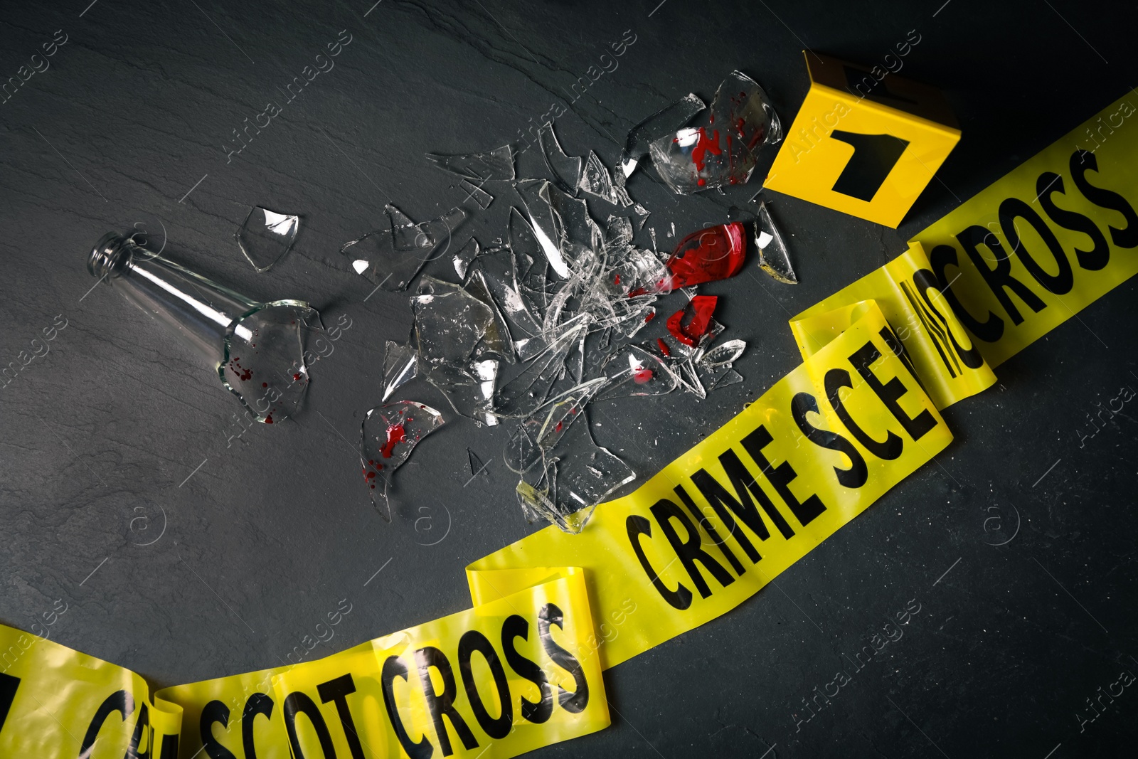 Photo of Broken bottle with blood, yellow tape and evidence marker on black slate background, flat lay. Crime scene