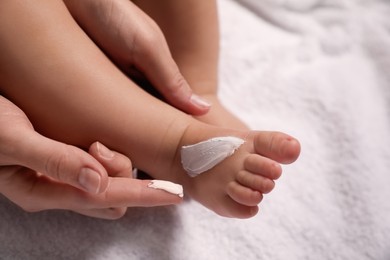 Photo of Mother applying moisturizing cream onto her little baby's skin on white towel, closeup