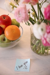 Paper note with handwritten text I Love you on white table near flowers. Romantic message