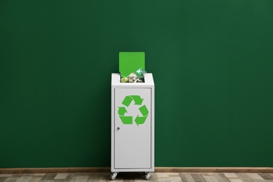 Overfilled trash bin with recycling symbol near color wall indoors