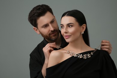 Man putting elegant necklace on beautiful woman against grey background