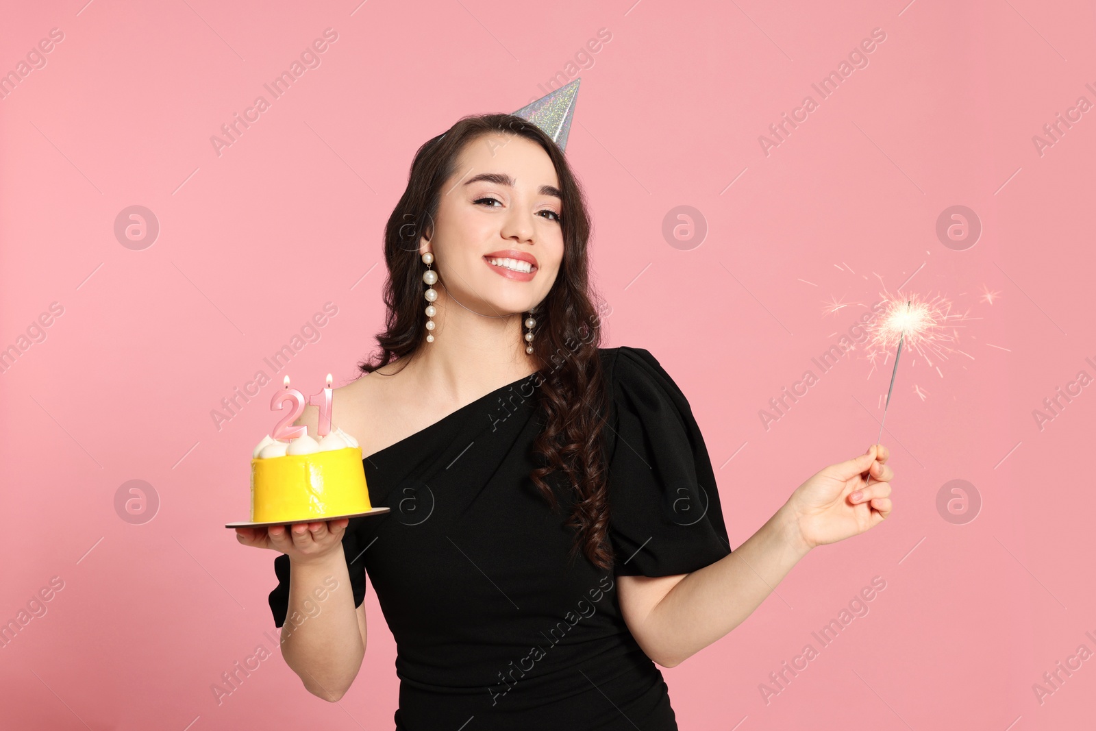 Photo of Coming of age party - 21st birthday. Smiling woman holding delicious cake with number shaped candles and sparkler on pink background