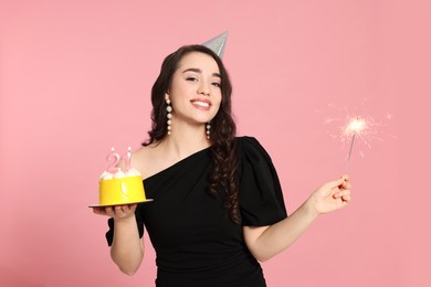 Coming of age party - 21st birthday. Smiling woman holding delicious cake with number shaped candles and sparkler on pink background