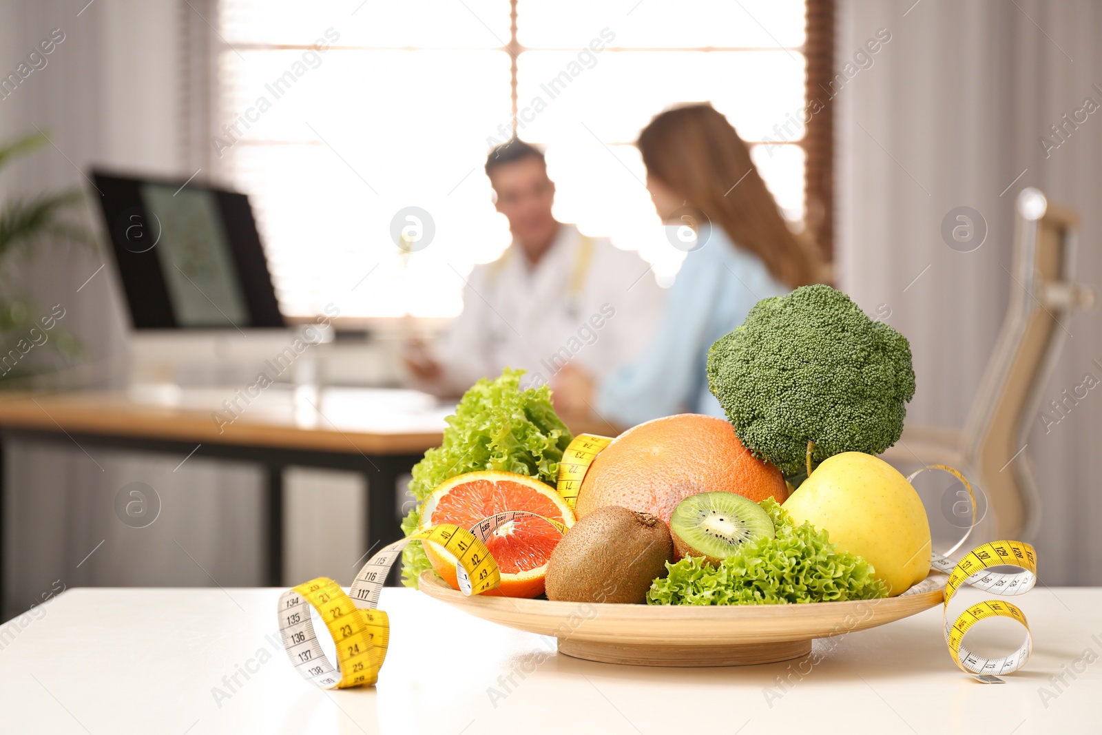 Photo of Nutritionist consulting patient at table in clinic, focus on plate with fruits, vegetables and measuring tape