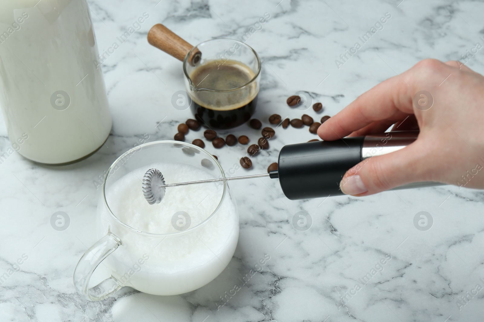 Photo of Woman whisking milk in cup with mini mixer (milk frother) at white marble table, closeup