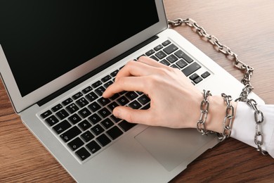 Photo of Woman with chained hand typing on laptop at wooden table, closeup. Internet addiction