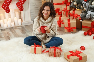 Image of Happy young woman holding cup of hot drink on floor with Christmas gifts
