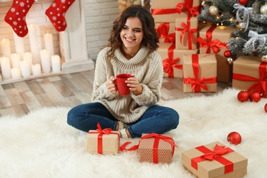 Happy young woman holding cup of hot drink on floor with Christmas gifts