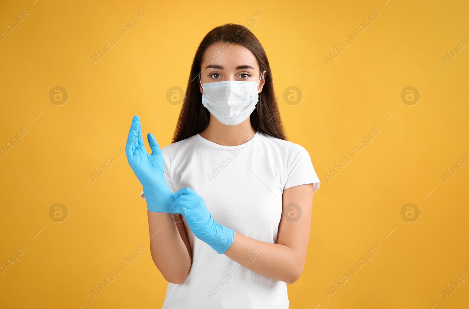 Photo of Woman in protective face mask putting on medical gloves against yellow background