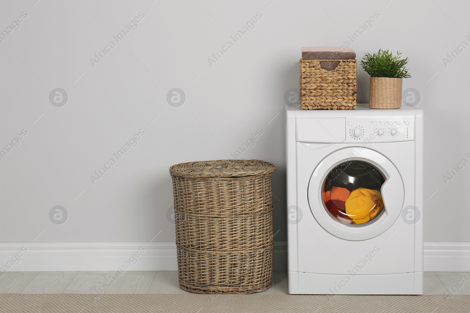 Photo of Modern washing machine with laundry, houseplant and wicker baskets near white wall