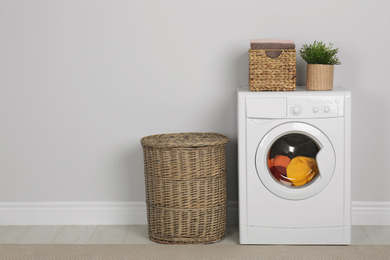 Modern washing machine with laundry, houseplant and wicker baskets near white wall