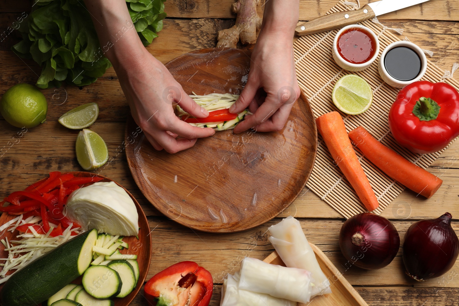 Photo of Making delicious spring rolls. Woman wrapping fresh vegetables into rice paper at wooden table, flat lay