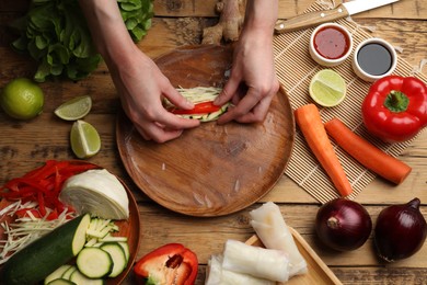 Making delicious spring rolls. Woman wrapping fresh vegetables into rice paper at wooden table, flat lay