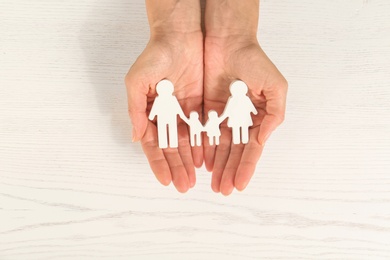 Photo of Woman holding figures of family in hands on white wooden background, top view