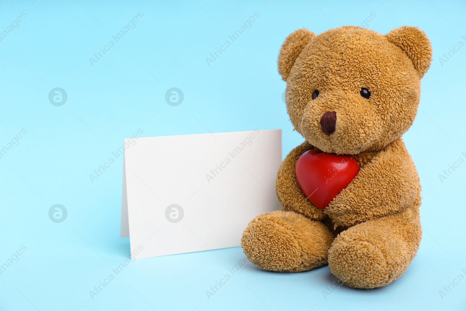 Photo of Cute teddy bear with red heart and blank card on light blue background. Valentine's day celebration