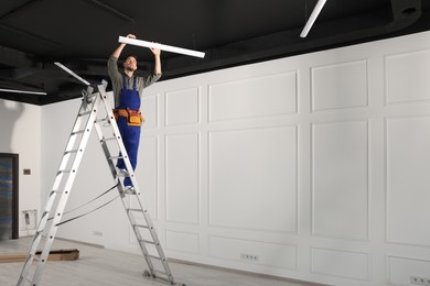 Electrician in uniform installing ceiling lamp indoors. Space for text