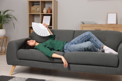 Young woman waving hand fan to cool herself on sofa at home