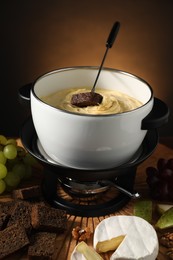 Photo of Fork with piece of bread, melted cheese in fondue pot and snacks on table against dark background, closeup