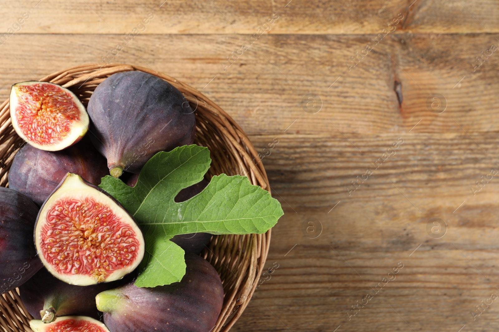 Photo of Wicker bowl with fresh ripe figs and green leaf on wooden table, top view. Space for text