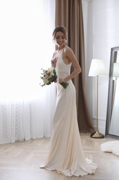 Bride in beautiful wedding dress with bouquet near window indoors