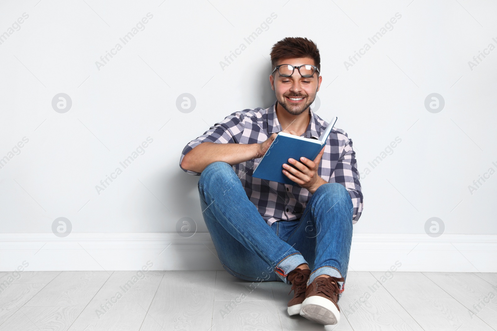 Photo of Handsome young man reading book on wooden floor near light wall