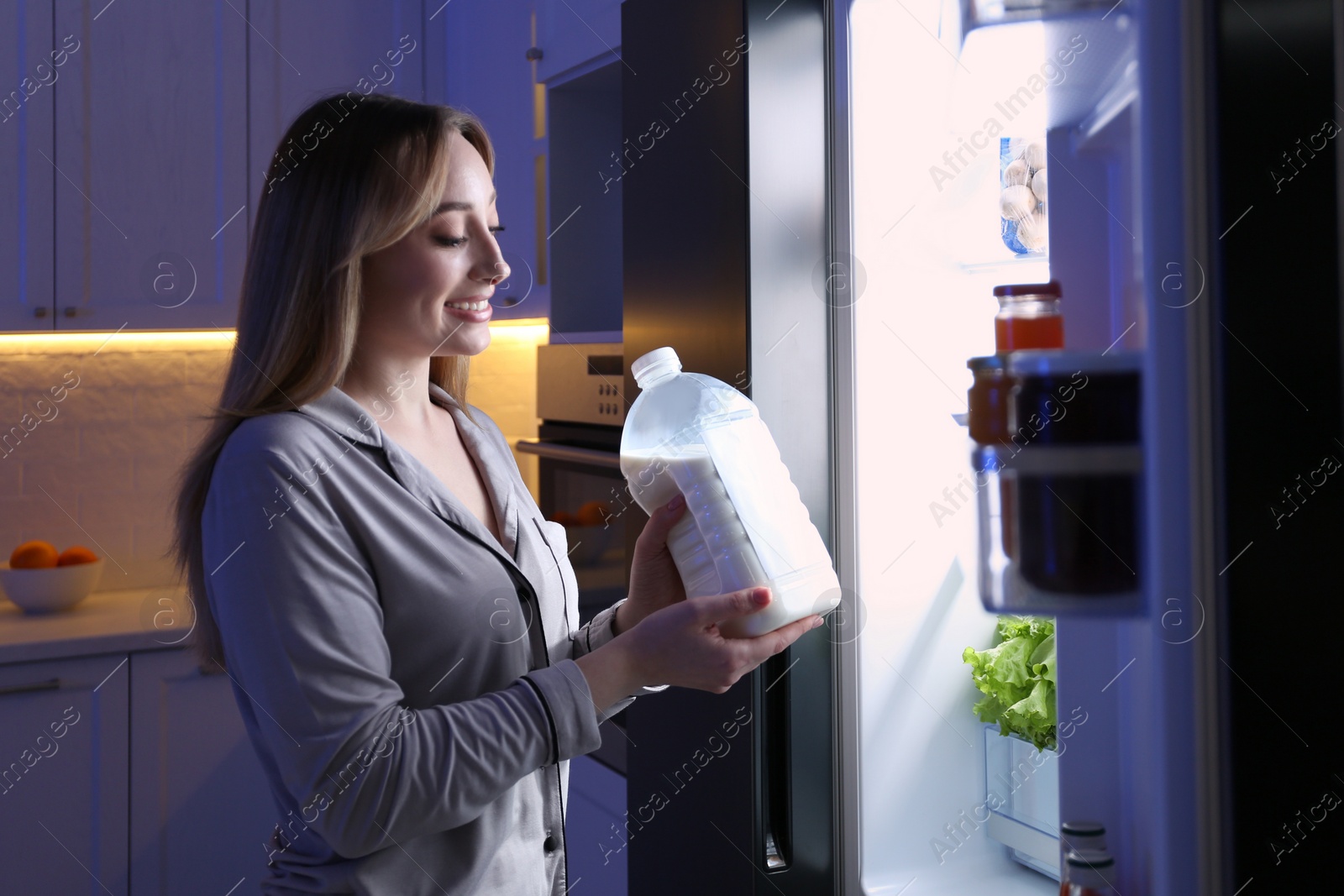 Photo of Young woman holding gallon bottle of milk near refrigerator in kitchen at night