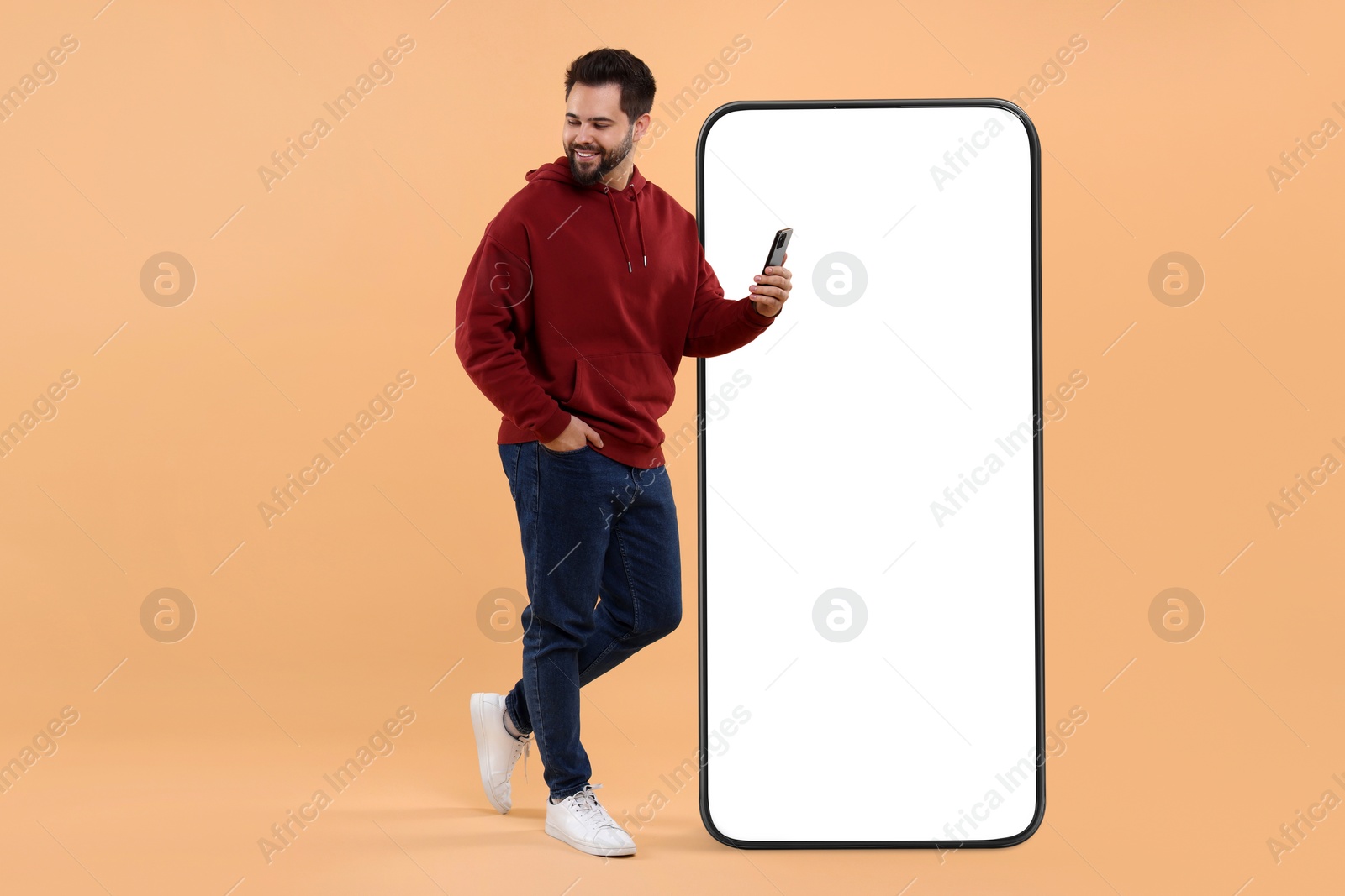 Image of Man with mobile phone standing near huge device with empty screen on dark beige background. Mockup for design