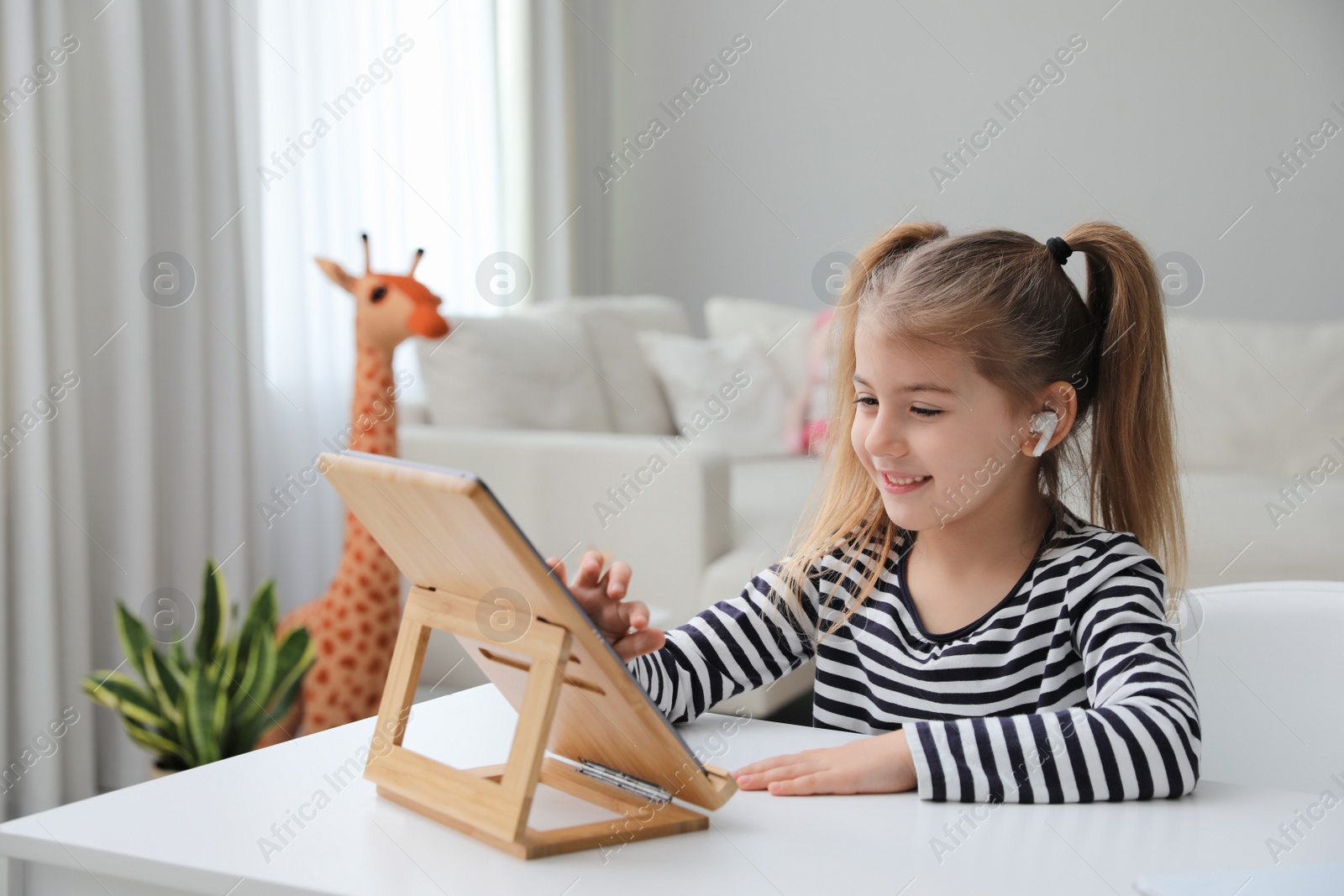 Photo of Adorable little girl doing homework with tablet and earphones at table indoors