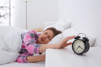 Photo of Sleepy young woman turning off alarm clock at home in morning, focus on hand