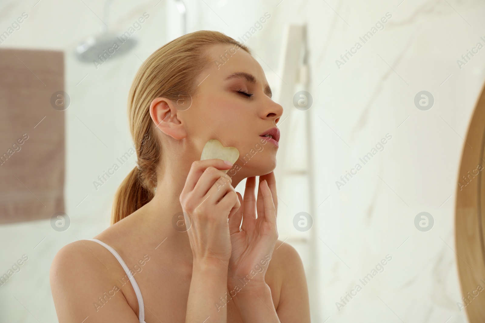 Photo of Beautiful young woman doing facial massage with gua sha tool in bathroom