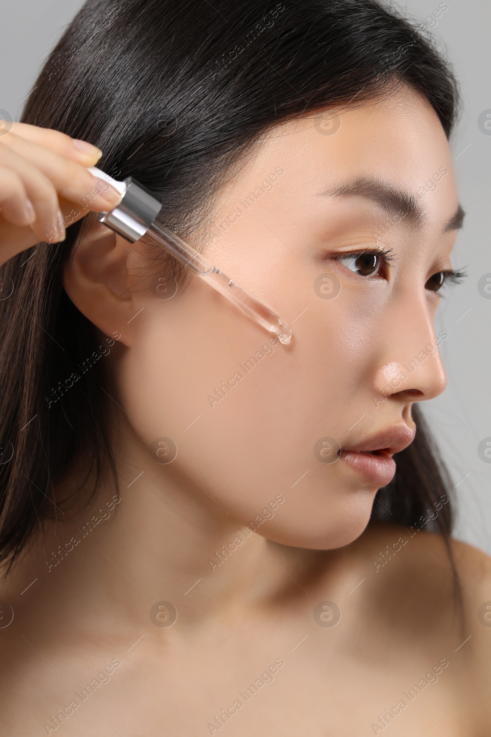 Photo of Beautiful young woman applying cosmetic serum onto her face on grey background, closeup
