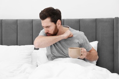 Photo of Sick man with cup of hot drink coughing on bed at home. Cold symptoms