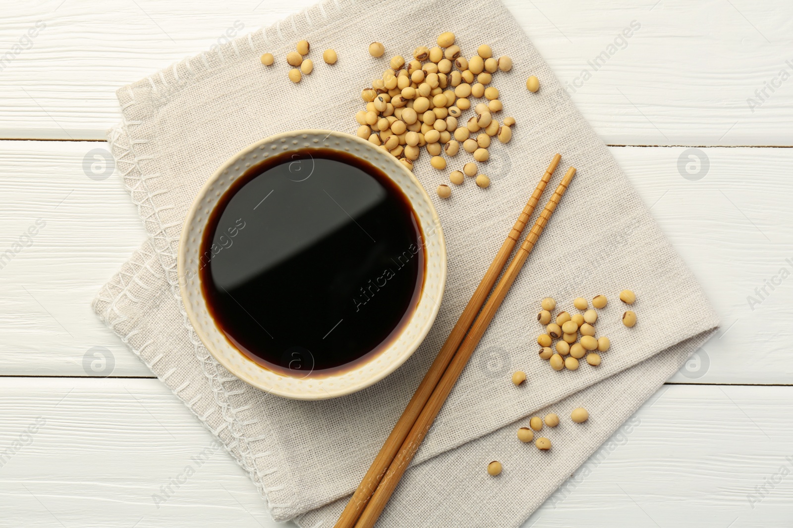 Photo of Tasty soy sauce in bowl, chopsticks and soybeans on white wooden table, top view