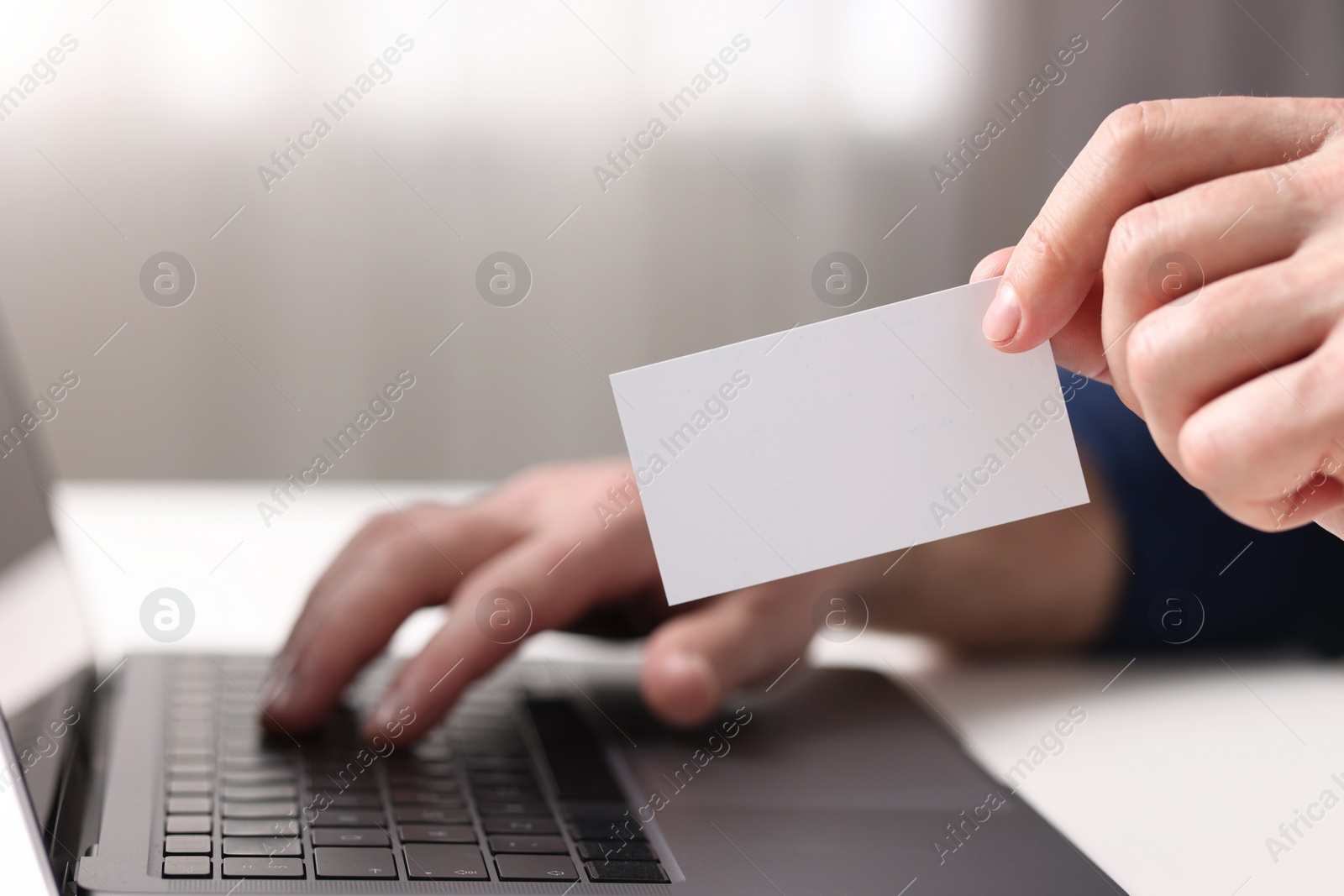 Photo of Man with laptop holding blank business card at white table indoors, closeup