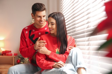 Photo of Happy couple in warm Christmas sweaters near window indoors