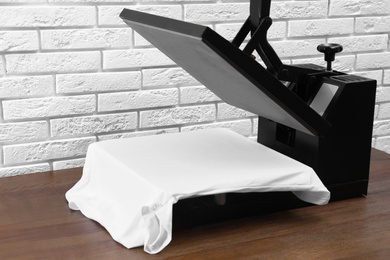 Photo of Heat press machine with t-shirt on wooden table near white brick wall
