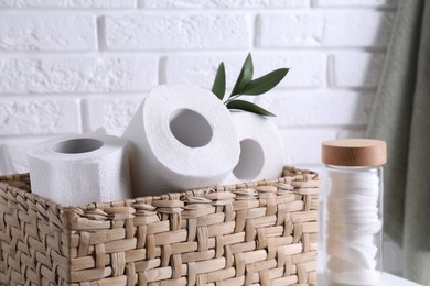 Photo of Toilet paper rolls in wicker basket, floral decor and cotton pads on table near white brick wall