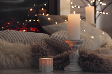 Burning candles, pillows and faux fur on floor near fireplace indoors. Bokeh effect