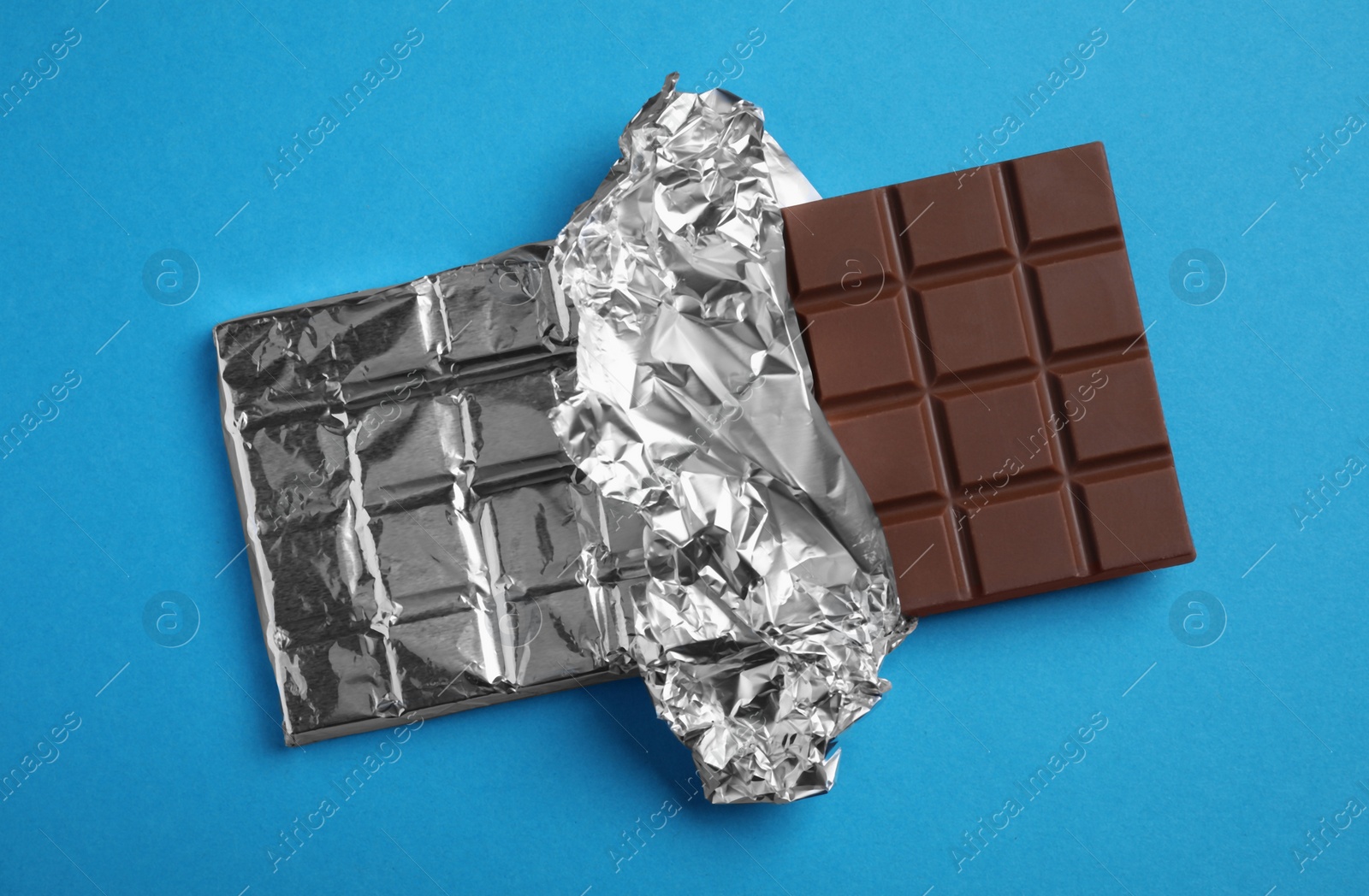 Photo of Delicious milk chocolate bar wrapped in foil on light blue background, top view