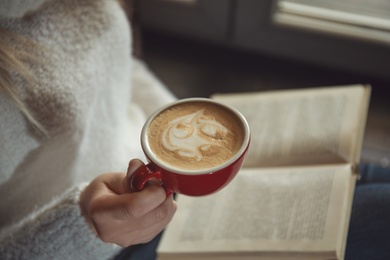Photo of Woman with cup of coffee reading book near window indoors, closeup