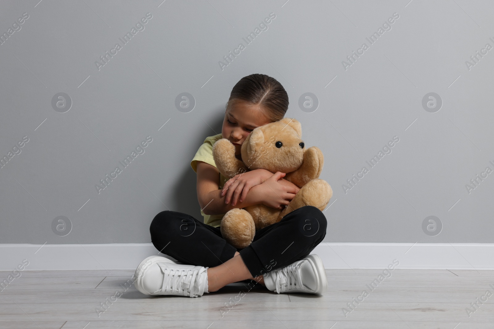 Photo of Child abuse. Upset girl with toy sitting on floor near grey wall