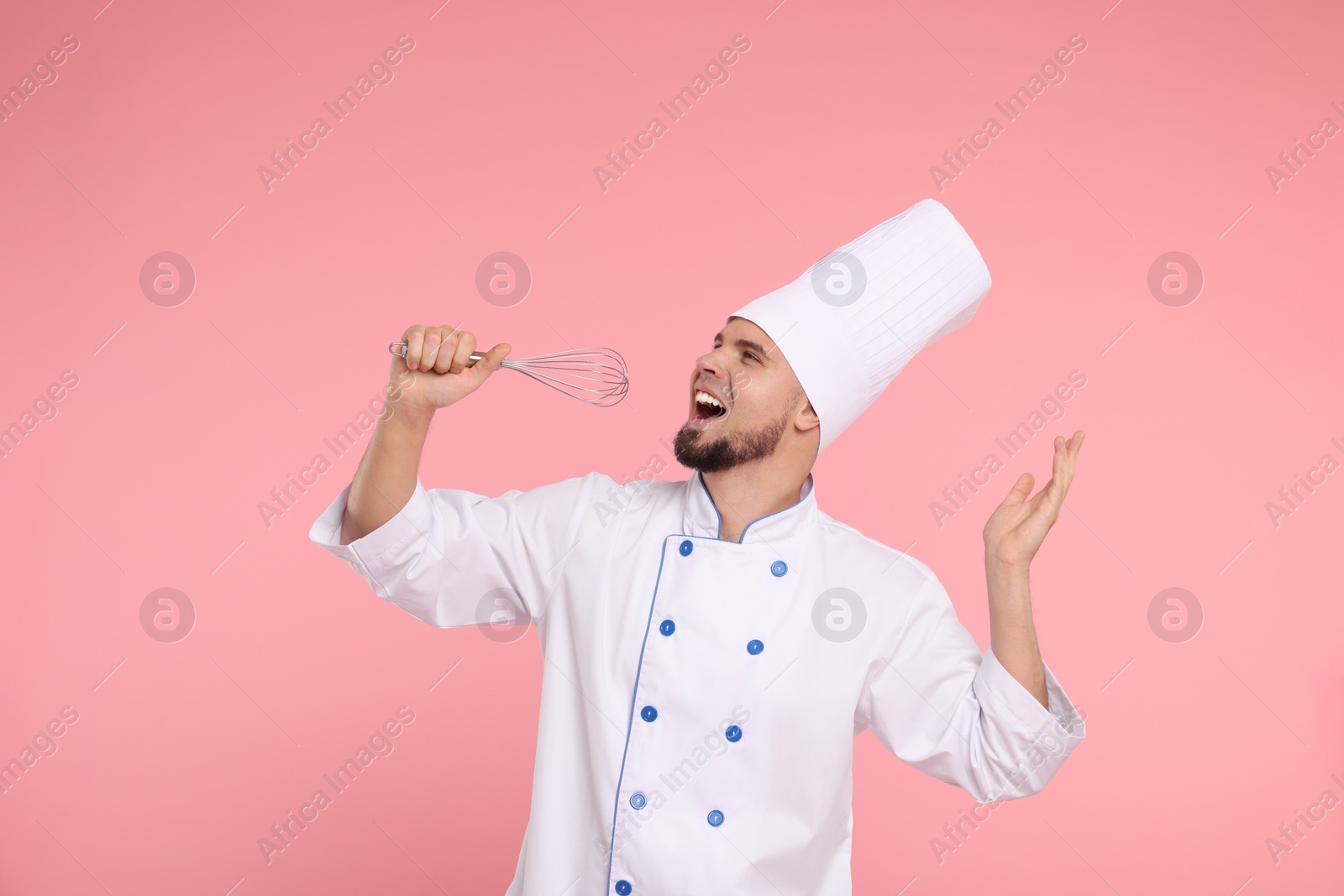 Photo of Happy professional confectioner in uniform having fun with whisk on pink background